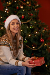 Happy woman, portrait and Christmas tree with gift box for present, giving or December holiday at home. Young female person with smile and hat for wrapped container, festive season or Santa at house