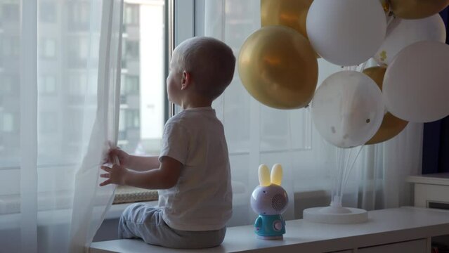 little boy sitting by the window with festive inflatable balloons, child looking out the window