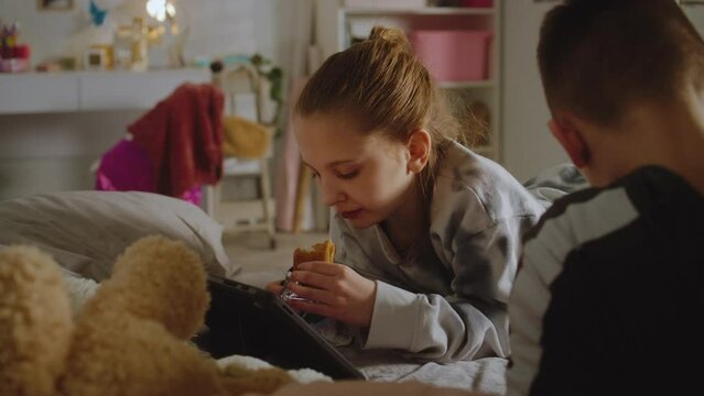 Young girl lies on bed, eats croissant, watches video content using tablet. Boy lies near sister and uses phone. Teenagers spend leisure time together. Family relationship. Home with stylish interior.