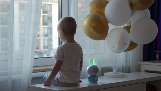 little boy sitting by the window with festive inflatable balloons, child looking out the window