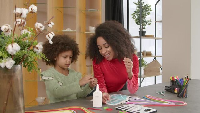 Creative engrossed in action cute preadolescent African American daughter with help of pretty caring mother making colorful paper quilling designs, rolling strips with slotted tool and talking indoors