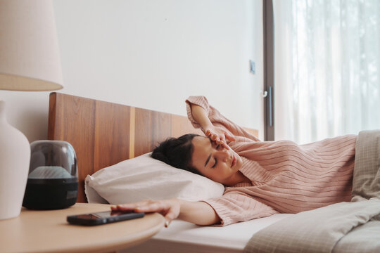 Young Asian woman in a soft pink sweater lying in bed with a headache, hand on forehead, with a serene expression and natural light pouring in from the window.