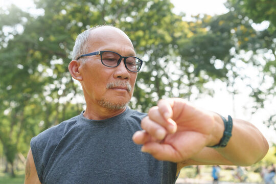 Asian senior man looking at smartwatch while exercising at park outdoor. Mature Adult male checking pulse after jogging. Fitness tracker