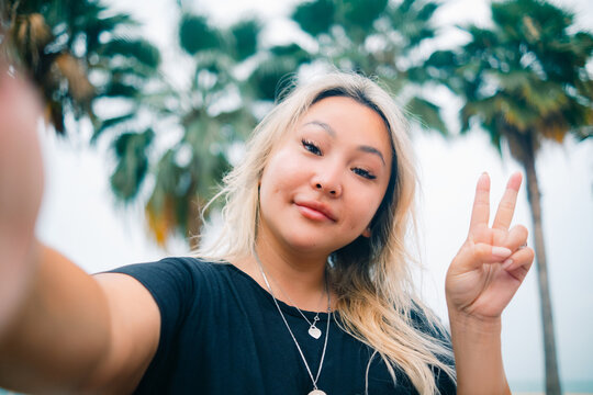 Young asian pretty woman shows V sign making selfie with happy smile with ocean beach palms on background