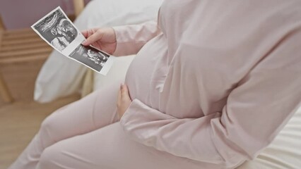 Hispanic pregnant woman holding ultrasound in bedroom, capturing expectancy and maternal joy indoors.