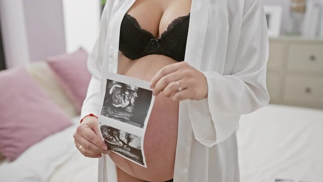 Pregnant woman holding ultrasound images, showcasing her belly in a home bedroom setting.