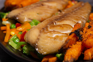 Smoked mackerel with vegetables and sweet potato fries on a black oval plate. On a dark stone background.