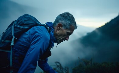 A man wearing a blue jacket and backpack is hiking up a mountain. The sky is cloudy and the man is looking up at the clouds - obrazy, fototapety, plakaty