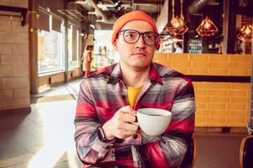A man with glasses and a orange hat enjoying a cup of coffee sitting in a coffee shop.
