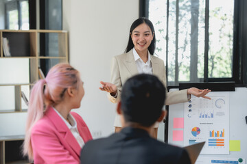 Asian businesswoman is giving a presentation to a group of people. She is smiling and pointing to a white board with graphs and numbers. The people in the audience are listening attentively