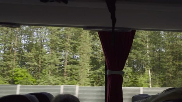 Side view from interior of tourist bus through window onto sunny roadway with noise barriers. Bus ride to distant suburb in forested landscape. Road trip in comfortable bus with passengers.