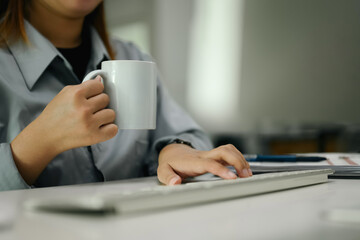 Cropped shot of female office worker holding coffee mug and typing on computer keyboard