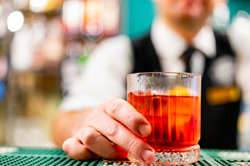 bartender serving a red cocktail at a bar. The focus is on the glass and the bartender’s hand. The drink is bright red and served in a short, clear, textured glass