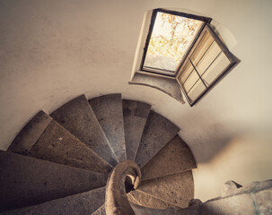 Old stone staircase in a tower - view from above at the stairwell. Perspective looking down view of clockwise round-shaped staircase. Small window overseeing the garden.