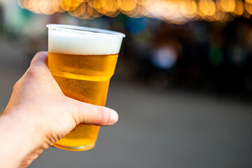 hand holding a cold beer in a plastic cup, set against a blurred festival background. The golden...