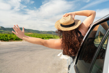 Rearview shot of an young woman leaning out of her car window with her arms outstretched on the road - obrazy, fototapety, plakaty