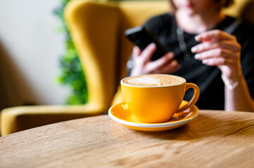 A bright yellow coffee cup with a creamy beverage sits on a wooden table. In the background, a blurred person uses a smartphone. The warm tones and artistic design make this scene inviting