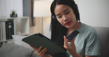 Selective focus, Happy young woman wear wireless headphones sitting on sofa using digital tablet and credit card for online shopping cashless in living room at home - 759606204