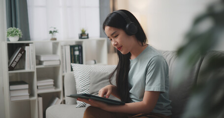 Selective focus, Young asian woman wear wireless headphones sitting on sofa using digital tablet for online shopping cashless in living room at home - 759606240