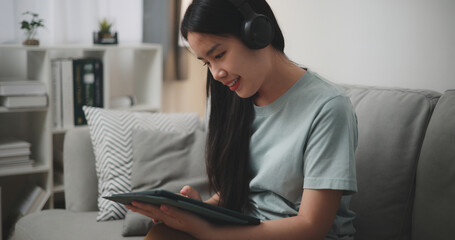 Selective focus, Young asian woman wear wireless headphones sitting on sofa using digital tablet for online shopping cashless in living room at home