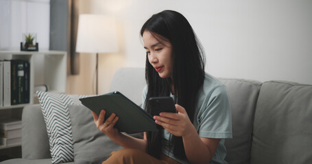 Selective focus, Happy young woman sitting on sofa using digital tablet and mobile phone for online shopping in living room at home - 759606499