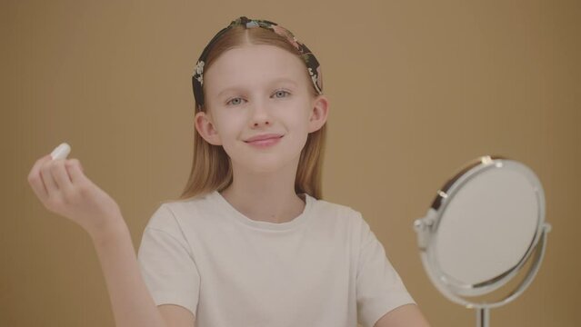 Zoom out effect. Funny blonde girl playing and having fun at home doing a beauty routine. Cheerful Caucasian teenage girl applies lip balm. Daily chapping routine. Beauty ads concept. Beige backdrop. 