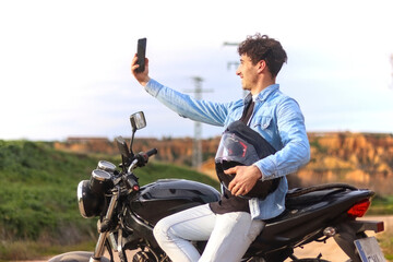 Young Caucasian man with curly hair sitting on his motorcycle taking a selfie smiling with landscape in the background. Lifestyle concept