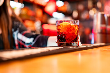 A close-up of a cocktail with an orange slice on a bar. The drink features a dark red liquid, and...