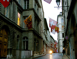 evening Geneva: quiet narrow European street; mid-rise buildings; Swiss flags; a lonely old man walks off into the distance; one policeman rides a motorcycle with his headlights on