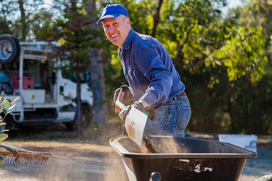 Happy smiling man shoveling dry dusty dirt into a wheelbarrow, gardening