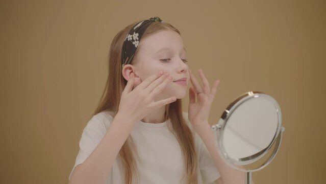 Nice youth girl putting face cream. Cute blonde girl looks in mirror and applies moisturizer on cheeks. Pure with glowing skin. Beauty and skincare concept for teens and influencers. Cosmetics Ad.