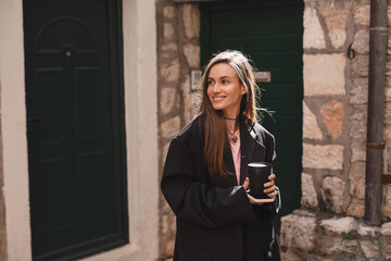 Happy beautiful stylish woman having good fashion clothes walking on street and holding coffee in cup takeaway to go with good mood. Girl wear black jacket look happy and look at side near green door.