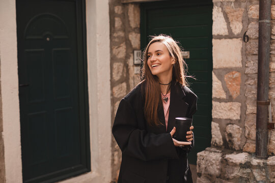 Happy beautiful stylish woman having good fashion clothes walking on street and holding coffee in cup takeaway to go with good mood. Girl wear black jacket look happy and look at side near green door.