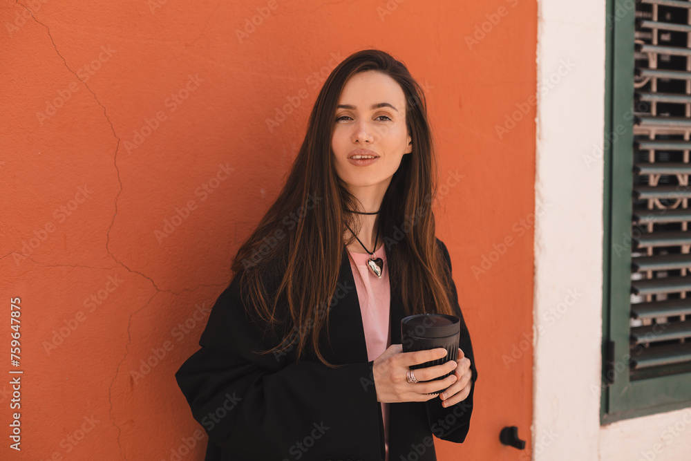 Poster Sensual woman standing near orange wall walking in old town, hold cup to go take away with coffee or tea. Woman drink cappuccino while rest on the street.  