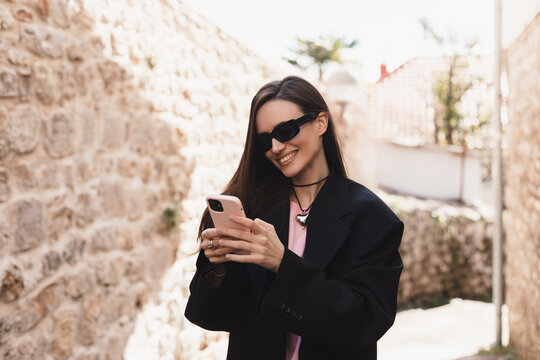 Photo of charming pretty young lady hold modern gadget smiling outside urban city street. Pensive brunette woman student freelancer wear black jacket walking in old town, hold phone in hand.