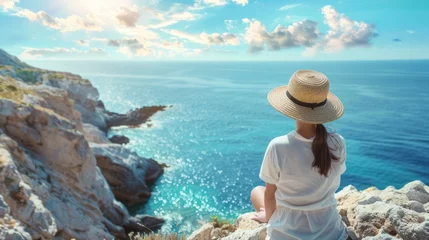Poster A young woman in a summer straw hat is sit on top of a cliff, looking at a sea view landscape with a blue sky. Travel concept for a couple or family road trip  © inthasone