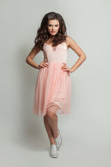 Elegant fashion model woman in pink dress and white shoes on feet standing against white studio wall background