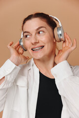 Modern woman smiling with braces on her teeth and listening to music in headphones on a beige background