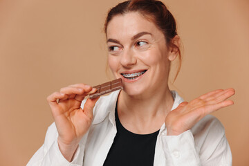 Portrait of mature woman in braces biting chocolate on a beige background