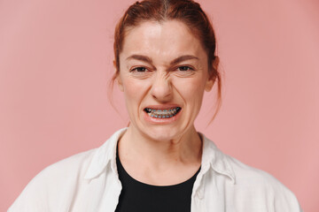 Portrait of red-haired mature woman suffering from pain due to braces on pink background