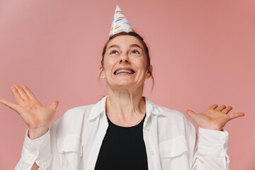 Portrait of smiling modern woman with braces and grimacing in birthday hat on pink background