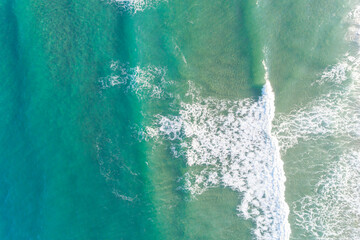 The ocean is calm and blue with white foam on the waves, aerial drone view