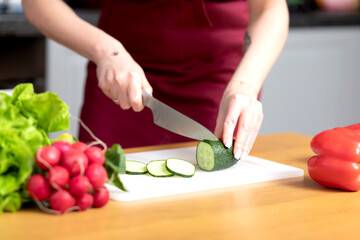 Housewife in red apron slicing cucumber and preparing vegetarian food	