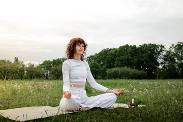 Concept of calmness. Sports woman sitting on yoga mat put hut on her lap. Female focused on breathing at the park. Full concentration mindfulness. Zen posture