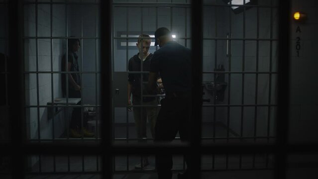 Warden takes off handcuffs from young prisoner standing behind metal bars. Multi ethnic teenagers serve imprisonment term in correctional facility or detention center. Guilty inmates in prison cells.