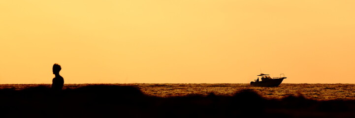 Silhouette of a young man walking on a beach at sunset, while a motor boat is going past. The scene...