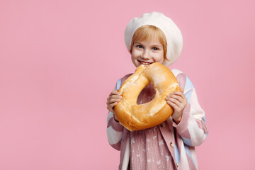 A Caucasian child in a white chef hat and striped apron holds a large golden bagel, pink...