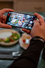 Hands holding a smartphone, capturing a vibrant spread of various dishes on a dining table in an indoor setting, possibly a restaurant.
