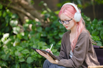 A cute pink-haired girl drawing on a digital tablet and wearing headphones in the garden, a Woman...