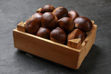 Roasted edible sweet chestnuts in wooden crate on grey textured table, closeup